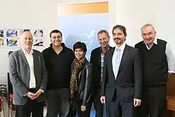 Shreen Abdul Saroor is pictured with other people who were awarded the Bremen Peace Award in 2011. Saroor is in the centre, surrounded by five other men. Saroor is wearing black pants, a leather jacket, and brown scarf. The men are dressed in business to casual attire, with multiple men in suits and one in a hoodie and jeans.