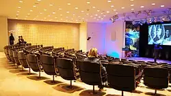 An auditorium in the basement, with black seats. The auditorium slopes slightly down toward a stage to the right.