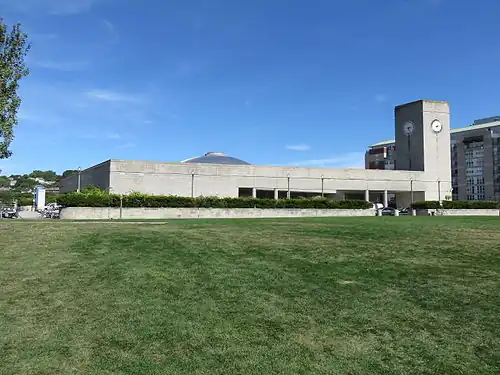 A low concrete building seen from across a lawn. It has a square clock tower at one corner and a low dome at its center.