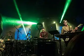 In this view of the band taken during the launch event for the The Race for Space album, J. Willgoose Esq., who performs on guitar and electronics, is pictured at right, and Wrigglesworth is pictured on drums at left. Live member JF Abraham is pictured at centre on bass guitar and electronics.
