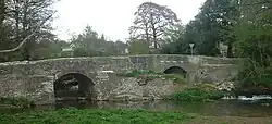 Gray stone bridge with two arches over water. The central pillar is on a small island. Trees to the left and right and behind the bridge.