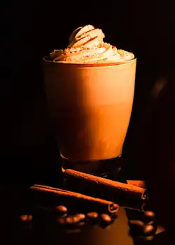 A pumpkin spice latte in front of a black background, in combination with two decorative cinnamon sticks and some coffee beans strewn in front.