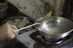 A cooked puri is removed from a pot of oil over a stove. Next to it are balls of uncooked dough.
