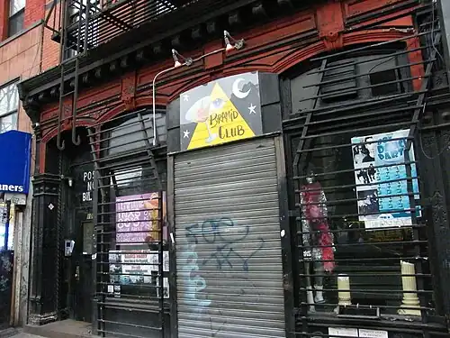 Exterior of the Pyramid Club, with a graffiti-marked metal shutter and a triangular sign featuring an eye, crescent moon, and stars above the door