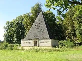Stone pyramid-shaped tomb with square base and decorated entrance, surrounded by grass and trees