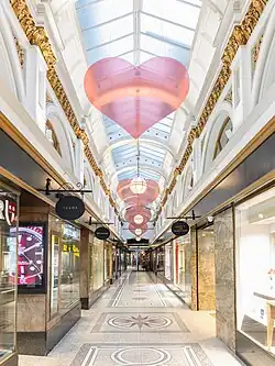 Photograph of the interior of Queen's Arcade in Belfast, taken in February 2026, showing luxury shops, walkways, decorative ceiling, and patterned floor tiles, highlighting the arcade as a high-end shopping destination.