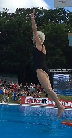 full-body action photo of Johanna Quaas jumping from a diving board into an outdoor in-ground professional swimming pool; the pool water appears blue because the pool walls are blue; there are trees in the far background, and a crowd of people is watching; Quaas is a tan Caucasian woman with short styled white hair and is wearing a black one piece bathing suit