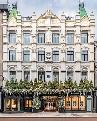 An ornate shopping arcade, lit up by ground floor shop windows, shop signs and fairy lights from above, in the tall ceiling space. The floor is surfaced with white tiles, with linear patterns along the edges of the walkway and diamonds interspersed along the centre.