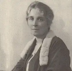 A seated woman, gray hair pinned up, in a black dress with fur collar before a plain white background