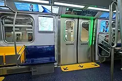 The interior of an R211 car, showing its LCD screens, digital advertising, yellow flip-up seats, door opening indicators, and looped stanchions