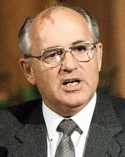 A man in a grey suit, white shirt and dark tie, balding with grey hair, he has a birthmark on his forehead