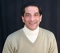 A man viewed from the chest up, wearing two sweaters at once. The outer layer is a beige v-neck knit sweater with long sleeves and a rib knit neckline. The inner layer is a white turtleneck sweater.