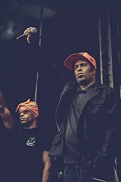 Lead Mano Brown standing on the right and supporting vocal Edy Rock standing on the left. Both are raising their right fists on the stage with a serious expression, Mano Brown holding the microphone.