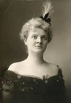 A white woman with blonde hair in a curly updo, wearing a black gown with an off-the-shoulder neckline