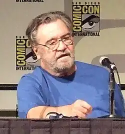Head and upper torso view of a lightly bearded, middle-aged man in glasses and a plain T-shirt, sitting behind a table with a microphone on it. Two small posters that read "Comic Con International" hang on the wall behind.