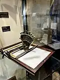 wooden frame holds a piece of a paper flat. On top of the paper is a brass device for making dots on the paper in the shape of Roman letters.