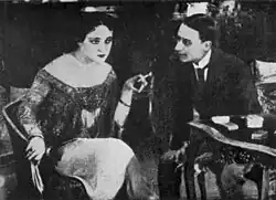 A woman in an elegant dress holds a cigarette while a man in a suit leans toward her in conversation