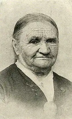 An older white woman with hair parted center and dressed to the nape; she is wearing a dark dress with a white underblouse or neck scarf