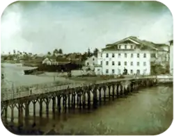 Photograph of a long wooden bridge crossing a river to a town with multi-storey white buildings and palm trees in the far background