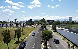Aerial photograph looking down a long highway with sparse trees on either side of the road, power lines, trees along the horizon and blue sky above it