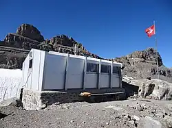 A white metal hut in the foreground, a mast with a Swiss flag to its right and rocky mountains in the background.