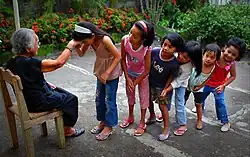 Six children lining up to their grandmother sitting on a chair. The one closest to the grandmother is holding up her hand to their forehead.