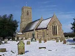Stone parish church with a square tower and flint walls, identified as St Margaret of Antioch in Reydon, Suffolk, viewed from the southeast. Snow covers the churchyard and roof, with scattered gravestones in the foreground and a war memorial statue near the entrance. The sky is bright blue with light clouds, and leafless trees frame the scene.