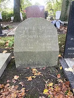 A gravestone with three names; the first and third are in Cyrillic; the dirt above is covered in autumnal leaves
