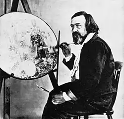 Photograph of a bearded, middle-aged man. He is seated and holding a paint brush. In front of him is an unfinished, circular painting on an artist's easel.