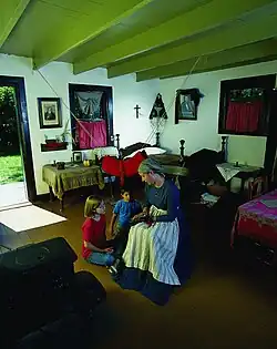 Interior view of Riel House showing a furnished common room with wooden walls, period furniture, and a wood-burning stove