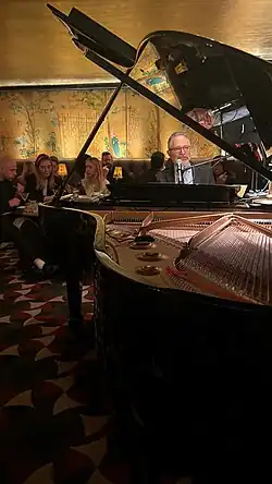 A man with a gray beard sits behind a grand piano, singing into the microphone. People at tables watch him intently. The carpet is lush and the backdrop is a yellowish wallpaper.