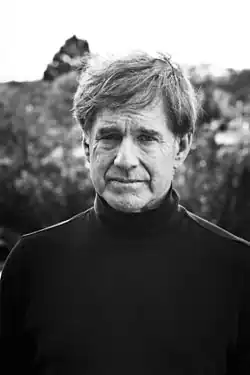 A black-and-white portrait of a man with tousled hair wearing a dark turtleneck, standing outdoors with a calm, serious expression