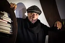 Ron Arad wearing dark clothing and a dark, striped hat, photographed leaning one arm against a bookshelf and the other against an unidentified object, in front of a white wall on which there is a shadow of the subject.