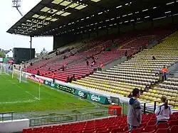 Part of a stadium, consisting of yellow and red seats. A grass football pitch is visible to the left.