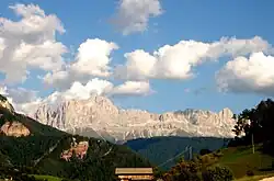 Photograph of a gray mountain chain. Closer to the viewer, there is a wooden house. Beyond there are green-tree-covered mounts. Further, a gray rocky steep treeless mountain chain. The sky is blue with big white clouds.