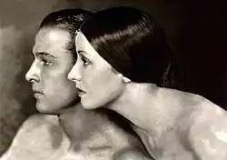 Man and woman posed in profile close together with smooth, slicked-back hair and bare shoulders in a soft-focus black-and-white portrait