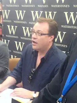 Davies in a navy-blue polo shirt, with one hand resting on a copy of his book.
