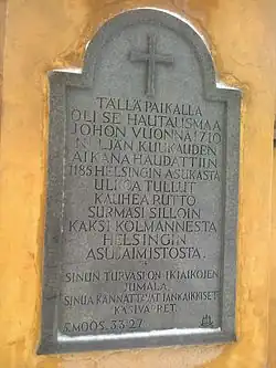 Stone commemorative plaque mounted on the gate of Old Church Park in Helsinki, bearing Finnish text about the 1710 plague burial ground and engraved with a cross at the top.