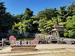 The Ryukyu Mura sign in the center of the village area, featuring the park's mascots: a kijimuna and a sentient taiko.