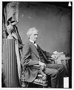 A well-dressed man in middle age facing sideways, sitting in a chair