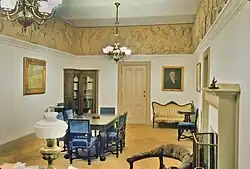 A grand office room, with painted portraits on the walls and ornate furniture. A strip of Greek sculpture, showing men riding horses, runs along the top of the walls.