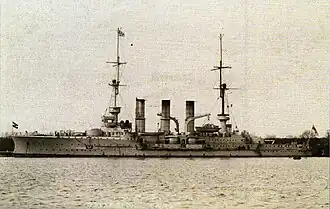 A large, light-colored warship with three tall funnels sits in port