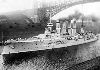 A large, light colored ship slowly passes through a narrow channel, underneath the arch of a bridge.