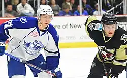 Ice hockey players in a Syracuse Crunch jersey and a Wilkes-Barre/Scranton Penguins jersey standing next to each other with their sticks being held down