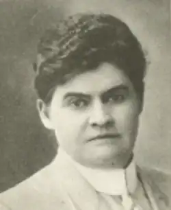 A woman with olive skin and short wavy dark hair, wearing a tailored jacket and collared shirt