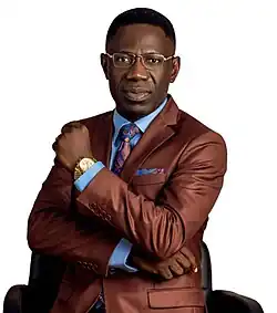 Colored profile image of Sam Oye wearing a brown suit, striking a pose with his left hand slightly raised, resting on his right hand