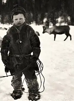 A child wearing a fur-lined winter jacket, thick trousers, and fur boots stands on snow, holding a rope, with reindeer in the background.