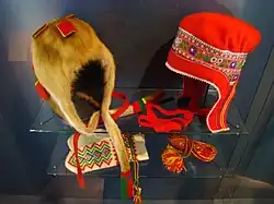 A glass-case display showing red fabric hats with decorative trim, a fur cap with tail, woven sashes, cords, and embroidered textiles.