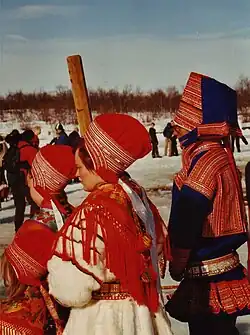 People wearing red and blue winter garments with patterned trim, tall fabric hats, wide belts, and shawls stand outdoors in snow during a spring gathering.