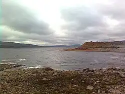 A body of water next to a rocky shore and hills under a cloudy sky.